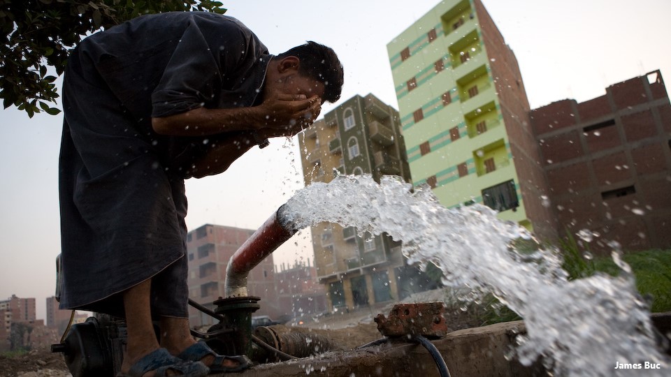 Water gushing from a standpipe by buildings, a man washing his face in the water.