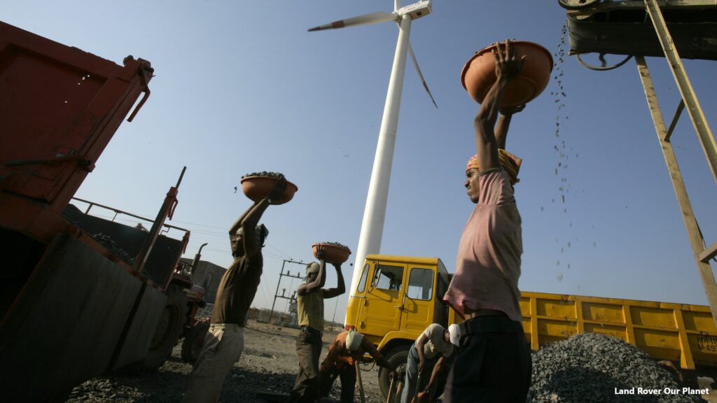 A laborer is seen working at diesel-powered crusher in front of a wind turbine. This is a 17.5 MW wind project, consisting of eighteen wind farm sites in the Indian states of Tamil Nadu, Maharashtra, Rajasthan and Karnataka. The eighteen sites have been bundled together for the purposes of carbon finance. The turbines displace electricity from the National Grid, which is dominated by coal and oil fired generation. The Indian electricity grid is dominated by fossil fuel powered generation