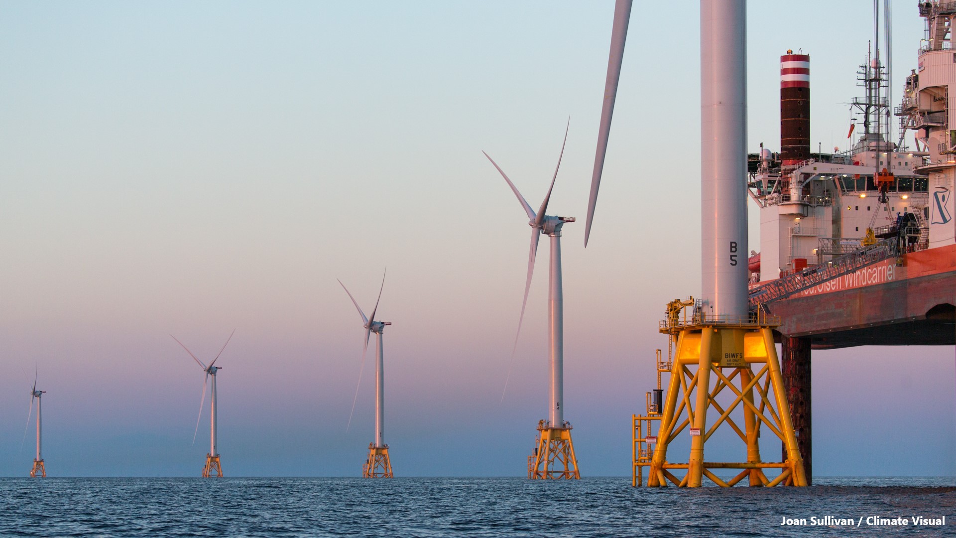 Dusk settles on the Block Island Wind Farm on the last day of construction - 18 August 2016 - of North America's first commercial offshore wind farm, developed by Deepwater Wind, now owned and operated by Ørsted. In this photo, the third blade has just been installed on the fifth and final GE-Alstom Haliade 150-6MW turbine by the Fred Olsen Windcarrier "Brave Tern" (partially seen on the right), which is in the process of jacking down to return to shore. The Block Island Wind Farm is located 3.3 nautical miles SE of Block Island, Rhode Island.