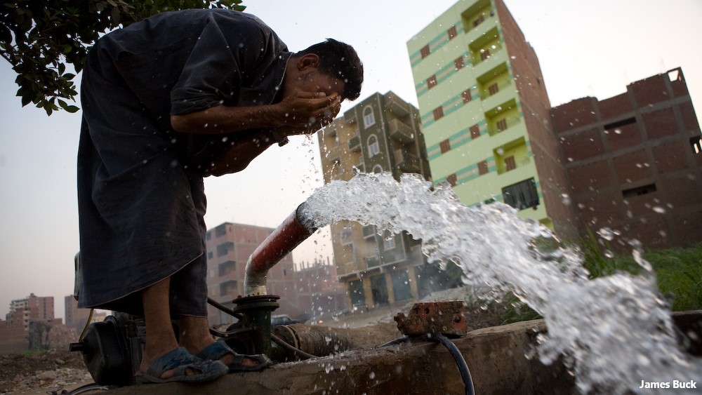 Water gushing from a standpipe by buildings, a man washing his face in the water.